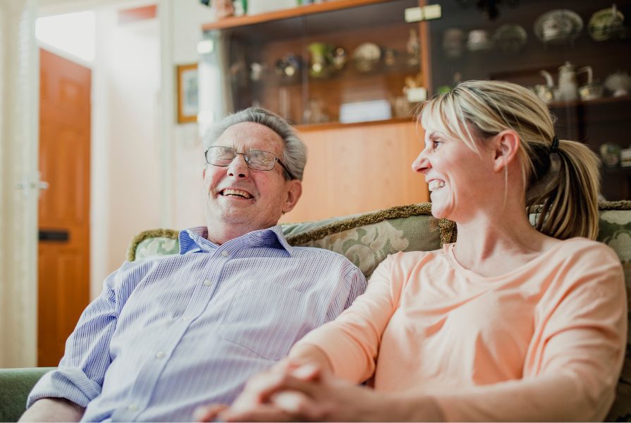 An elderly white man laughing sitting next to a middle aged white woman on a sofa who is smiling at him.